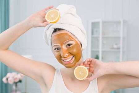 Beauty Skin Care Concept. Attractive Caucasian woman in white towel with brown facial mask on face holds citrus fruit on her hand on light background. Spa procedures and cream mask on skin.の写真素材