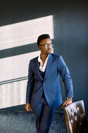 Young beautiful African American guy in a stylish suit standing in the office laying his hand on a chairの写真素材