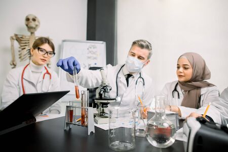 Medicine, genetics, immunology, microbiology concept. Multiethnic group of scientists, Caucasian man and woman, Muslim woman, working at the laboratory with microscope, tubes with liquids and laptop.の写真素材