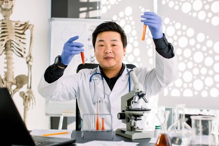 Smiling Asian man scientist or doctor testing new drugs to prevent and destroy new strains of the virus or bacteria, holds two glass tubes. Pharmaceutical research and medicine concept.の写真素材
