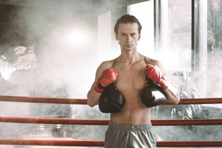Serious Young muscular man boxer in boxing ring. Boxing gloves slung over his shoulder on dark background indoorの写真素材