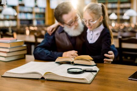 Grandfather and granddaughter reading a book in old vintage city library. Family reading, leisure, education concept. Focus on books and magnifying glass on the tableの写真素材