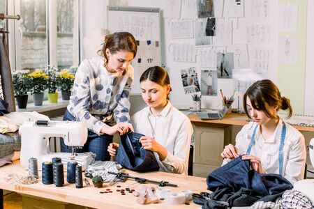 Young smiling fashion designer posing with scissors at working place and looking at camera while two pretty dressmakers sitting at the table and sewing in working process.の写真素材