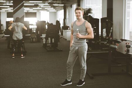 Resting time. Smiling young man in sportswear standing with bottle of water and looking at camera on the background of modern fitness gym.の写真素材
