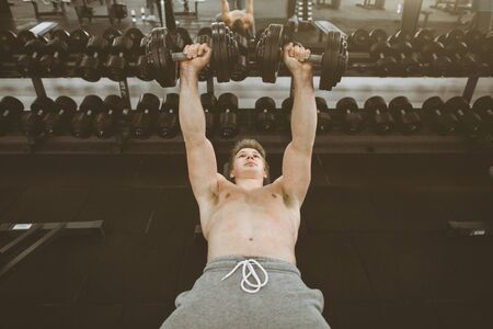 Handsome young phisique fitness man works out in gym lifting dumbbells. Man bodybuilder exercising at gym holding dumbbellsの写真素材