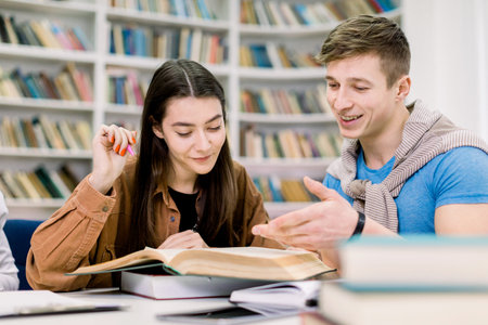 Students, Caucasian handsome boy and pretty girl, sitting at a table in a library while reading...