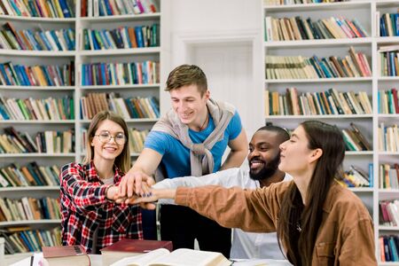 Exams and project preparation. Multiracial college students putting their hands on top of each other symbolizing unity and teamwork, sitting together in libraryの写真素材