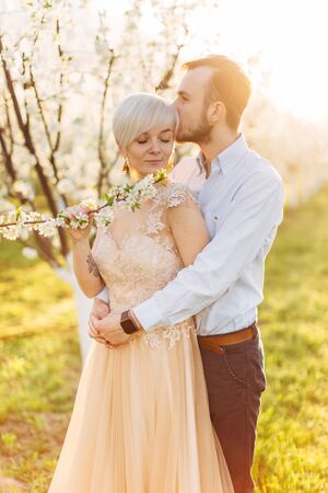 Man and woman in blooming garden on spring day. Handsome bearded man hugging and kissing his beautiful charming woman in light orange elegant dress. Passion and love conceptの写真素材