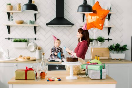 beautiful mother and little daughter at the celebration of a childs birthday in a light decorated kitchen. Mother and baby in birthday hats having fun, focus on the table with cake and presentsの写真素材