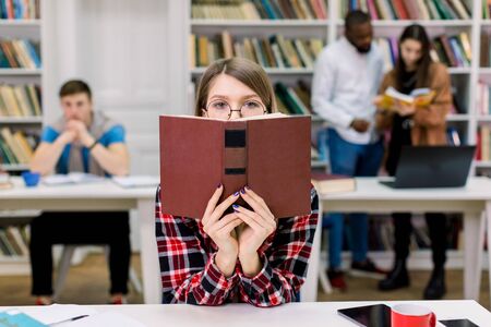 High school, education and people concept. Pretty student girl hiding her face over open book on modern college library background and multiethnic friends studying togetherの写真素材