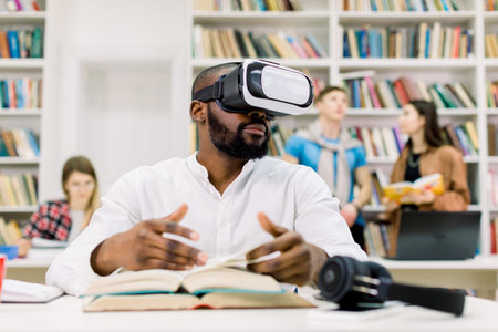 Young attractive African man with beard wearing casual white shirt, studying in library, using...