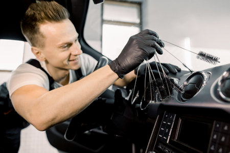 Young handsome smiling Caucasian man in black protective gloves using cleaning brush and removing dust from car control panel. Car detailing or valeting concept. Focus on handの写真素材