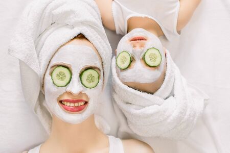 Mother and daughter doing funny spa procedures after bath. They are in white bath towels with white facial mud mask on faces and slices of fresh cucumber on their eyes. Family spa, mothers dayの写真素材