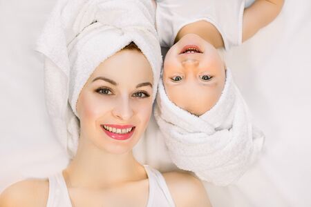 Smiling mother and daughter lying on white background, bathtime concept. Pretty young woman with little girl wearing towels on head after shower lying down and looking at camera on isolated backgroundの写真素材
