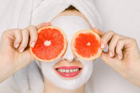 Pretty young girl in bath towel and facial mask with grapefruit slices in front of her eyes. Beautiful woman holding grapefruit slices like eyeglasses, hiding her eyes, lying on white backgroundの写真素材