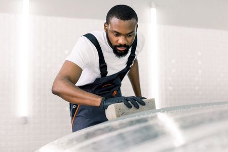 Young car service African male worker in black rubber gloves amd gray overalls, doing cleaning the...