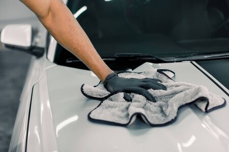 Car detailing, wash and cleaning concept. Cropped image of hand of male professional car wash worker in black rubber gloves, holding the gray microfiber and polishing the car hood of luxury white car.の写真素材