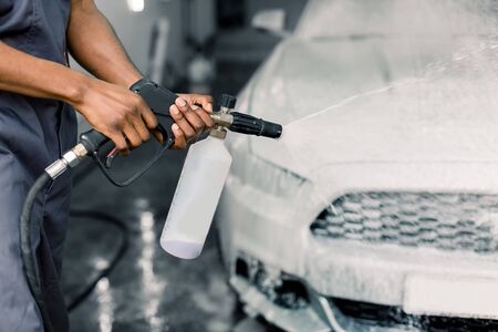 Manual car washing concept. Cropped image of hands of young dark skinned man worker cleaning modern white car with foam and pressured water at service station. Manual detailing car washの写真素材