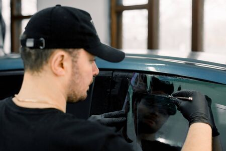 Back angle view of car detailing workshop male worker, wearing black cap and t-shirt, applying tinting foil on a car window in a garageの写真素材