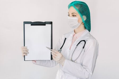 Young woman doctor holding empty blank and pen in hands. Female doctor wearing white coat, stethoscope, medical face mask and cap, showing a clipboard with blank paper. Isolated on white backgroundの写真素材