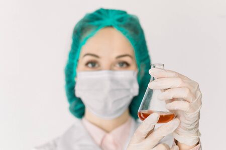 Concept of healthcare and medicine, COVID-19 . Serious female doctor virologist holding in hands glass chemical flask with red liquid. Development of new drug or vaccine. Focus on flask in handsの写真素材