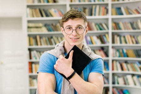 young smiling student in blue t-shirt and eyeglasses, holding black book in hands, and looking at...