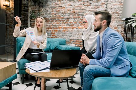 Group of three focused multiethnic male and female business people in formal clothes gathered around laptop on the table in modern office and disscussing key stages of business projectの写真素材