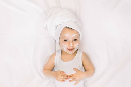 Little child baby girl having fun after bath with towel on head and face cream on cheeks, nose and forehead, lying on white background. Baby hygiene, face care, spa and bathの写真素材