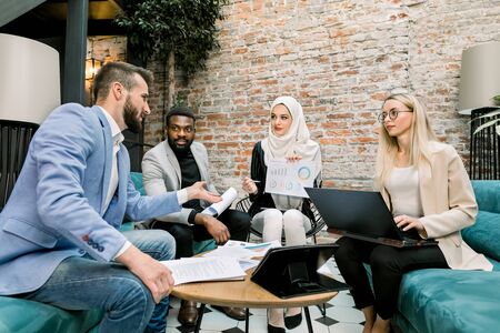 Multiethnic business people on the meeting in office. Arabian business lady in white hijab using financial document with graphs talking to her male and female partners about the results of researchの写真素材