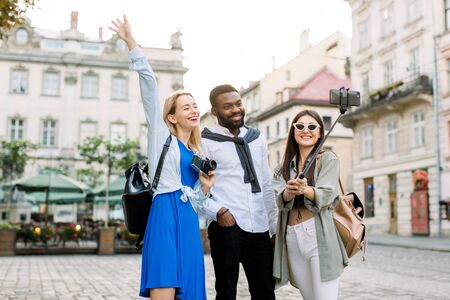 Excited cheerful mix raced friends enjoying walking outdoors in European city. Man and two women walking outside, hugging each other, laughing and making selfie photo. Multicultural friendship conceptの写真素材