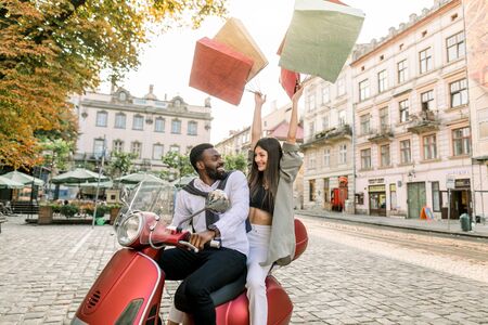 Charming smiling Caucasian woman sitting behind her handsome African boyfriend on the red scooter with shop bags in her hands raised up on the background of European city buildingsの写真素材