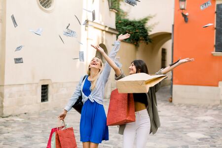 Two pretty young Caucasian women, wearing summer casual clothes, walking together with shopping bags and throwing paper money in the air. Shopping in the city, women friendship conceptの写真素材