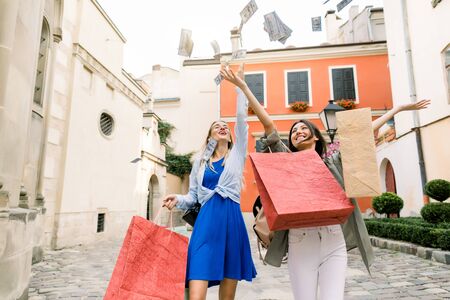 Two pretty young Caucasian women, wearing summer casual clothes, walking together with shopping bags and throwing paper money in the air. Shopping in the city, women friendship conceptの写真素材