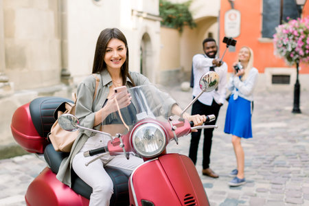 Pretty cheerful brunette Caucasian girl sitting on the red scooter in old city street, while her...