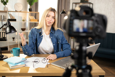 Happy blond caucasian woman in jeans shirt recording online tutorial on modern camera. Smiling female using laptop and camera during remote work at home.の写真素材