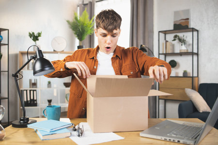 Excited surprised young Caucasian man opening box with ordered goods while sitting at the table at...