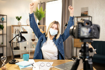 Happy woman in face mask talking on camera that fixed on tripod. Young blogger sitting at table with...