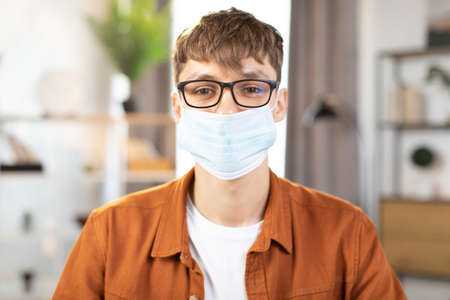 Happy Caucasian man in face mask and in casual wear sitting at table with modern laptop, making notes and smiling on camera. Concept of people, technology and distance learning.の写真素材