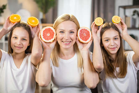 Portrait of happy mother with two pretty daughters smiling on camera and holding slices of grapefruit, lemon and oranges in hands. Three women using fruits for beauty procedures at home.の写真素材