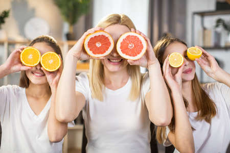 Happy sisters and their charming mother covering eyes under slices of grapefruit, lemon and orange while sitting at living room. Time for beauty procedures at home.の写真素材