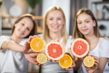 Blur view of woman and two teen girls posing on camera with slices of citrus in hands. Caring mother and two daughters enjoying spending time for beauty rituals at home. Selective focus on fruitsの写真素材