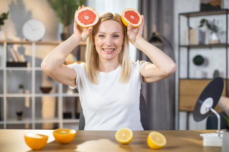 Smiling likable middle aged woman holding slices of grapefruit near her face while smiling on...