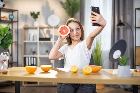 Domestic lifestyles of pretty girl. Youth and natural beauty concept. Pretty teen female holding slice of grapefruit and smiling while taking selfie on modern smartphone.の写真素材