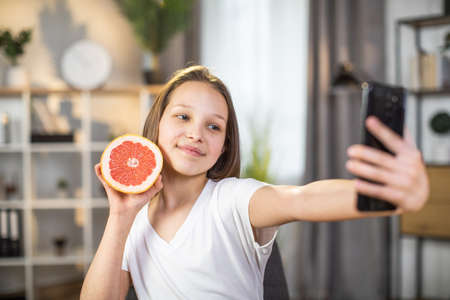 Pretty caucasian girl using modern smartphone for taking selfie with slice of grapefruit in hand. Female teenager with clear skin on face using citrus for beauty care at home.の写真素材
