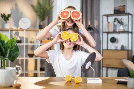 Two happy sisters covering eyes with slices of grapefruit and orange while posing on camera at bright living room. Fresh and pure skin of pretty teenagers. Youth and beauty concept.の写真素材