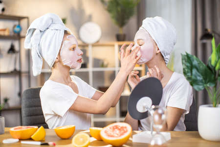 Two pretty sisters with towel on head applying mask on face while sitting at desk. Young caucasian females doing beauty treatments together at home.の写真素材