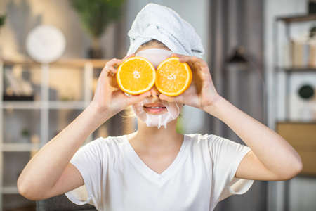 Close up portrait of pretty female teenger with towel on head and mask on face covering eyes with slices of oranges. Mineral and vitamin cosmetic for skin care at home.の写真素材