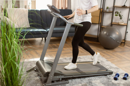 Cropped image of girl running on treadmill with a bottle of water in hand in modern bright...