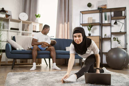 Arab woman in headscarf on her head doing gymnastics in the living room at home while african husband sitting on sofa with multinational little son in his arms. Enjoying Healthy Lifestyle, Copy Spaceの写真素材