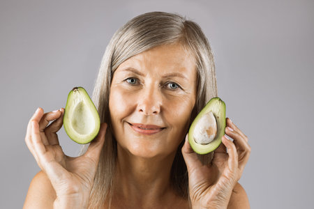 Portrait of pleasant mature woman posing in studio with halves of green ripe avocado in hands....
